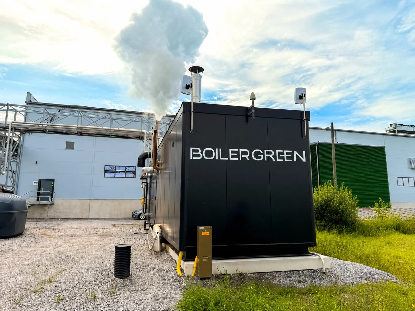 A black BoilerGreen hybrid boiler container beside a factory in summer, with steam rising from its stack